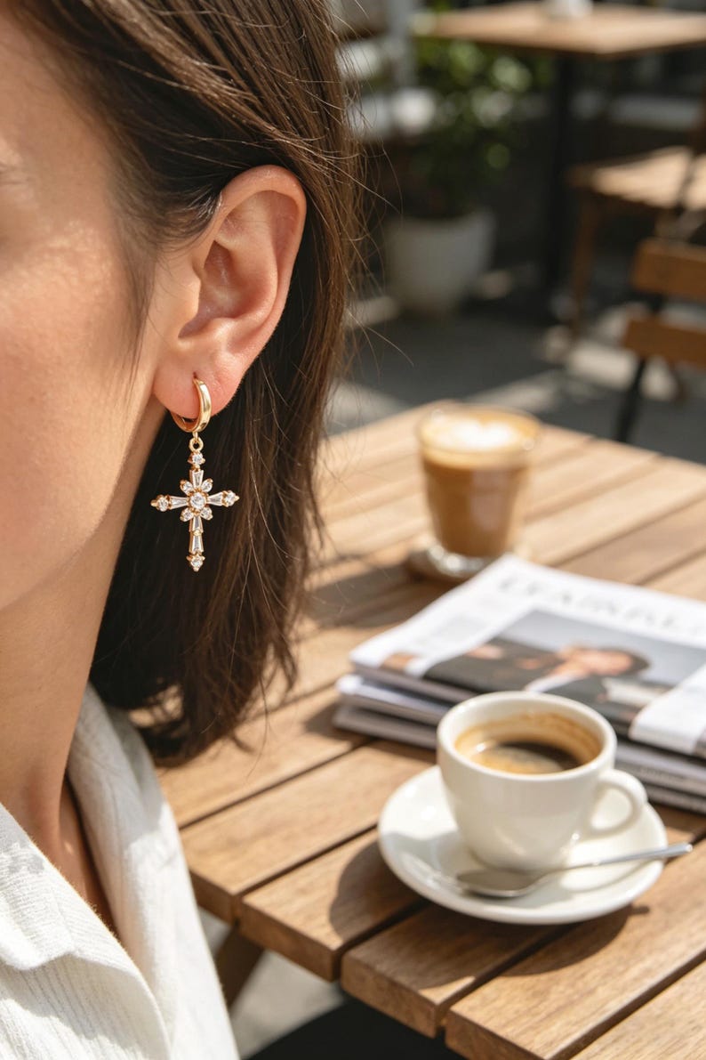 Pu&ograve; includere: Orecchini a croce color oro con pietre preziose chiare e scintillanti. Gli orecchini sono appesi a un cerchio d'oro. L'immagine mostra un primo piano degli orecchini, con uno sfondo sfocato di un caff&egrave;.