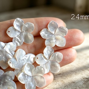 May include: A collection of white, iridescent, five-petaled flower charms carved from shell. Each floral piece features a small hole for attachment and measures 24mm in diameter. The surface of the charms displays a natural, pearlescent sheen.