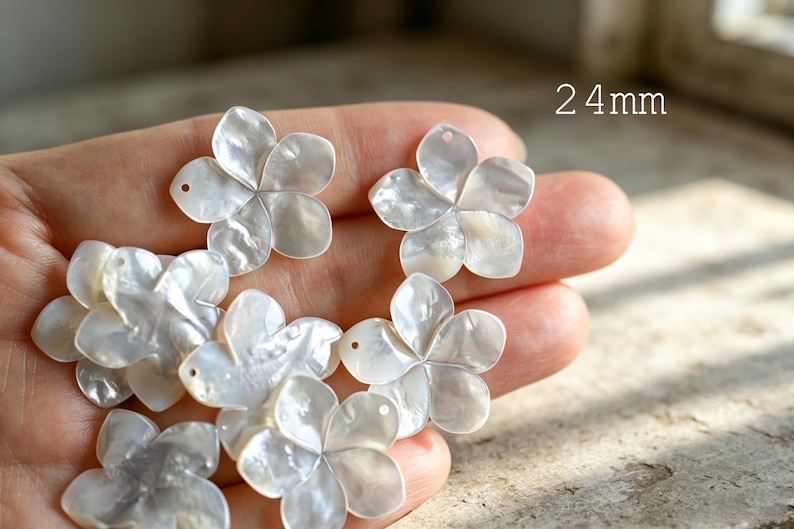 May include: A collection of white, iridescent, five-petaled flower charms carved from shell. Each floral piece features a small hole for attachment and measures 24mm in diameter. The surface of the charms displays a natural, pearlescent sheen.