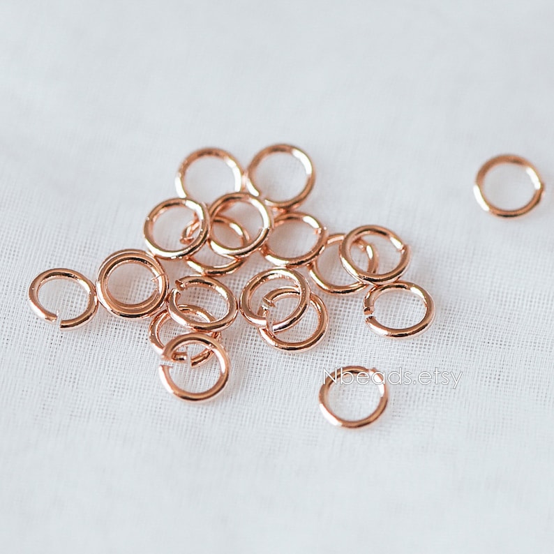 May include: A pile of rose gold jump rings, a type of jewelry finding, on a white fabric background.