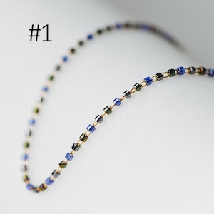 May include: A delicate chain necklace made of small, square-shaped beads in shades of blue and black. The beads are strung on a thin gold wire.