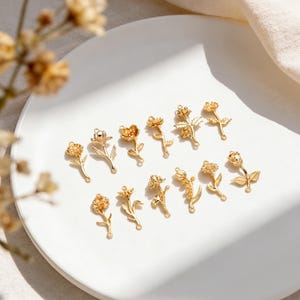 May include: A collection of gold-colored floral charms, each with a unique flower design. The charms are arranged on a white plate, showcasing their intricate details and delicate stems. These charms are ideal for jewelry making or craft projects.