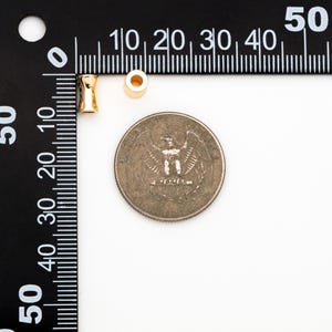 May include: Two gold-colored metal beads, one round and one cylindrical, next to a US quarter for size comparison.