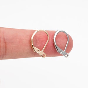 May include: Two earring hooks, one gold and one silver, are shown on a finger. The hooks are made of metal and have a simple design.