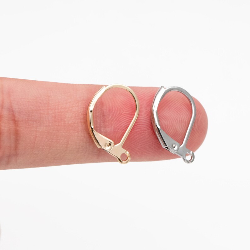 May include: Two earring hooks, one gold and one silver, are shown on a finger. The hooks are made of metal and have a simple design.