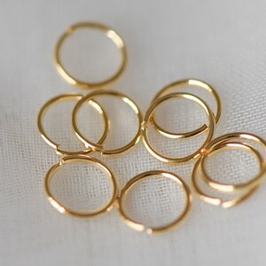 May include: A group of eight gold jump rings, arranged in a loose pile on a white fabric background.