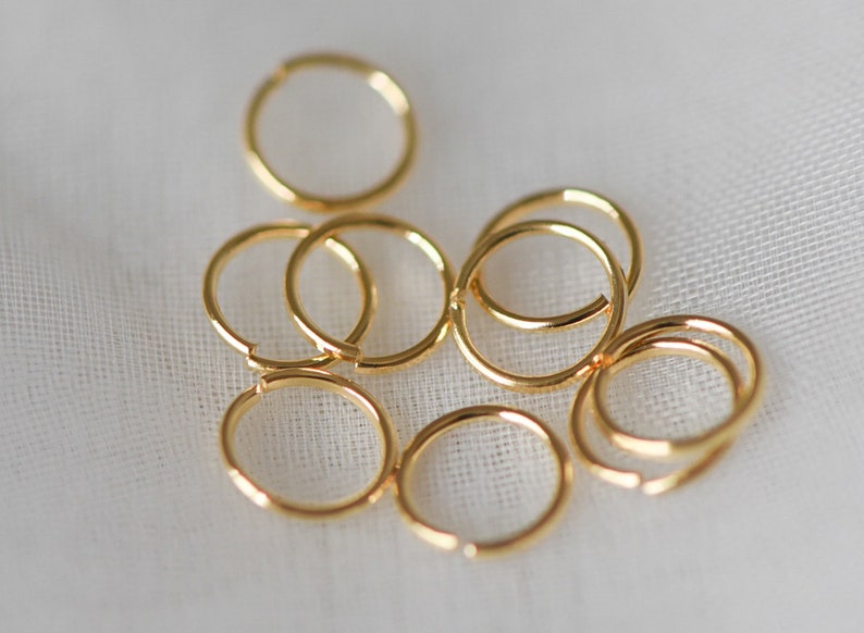 May include: A group of eight gold jump rings, arranged in a loose pile on a white fabric background.