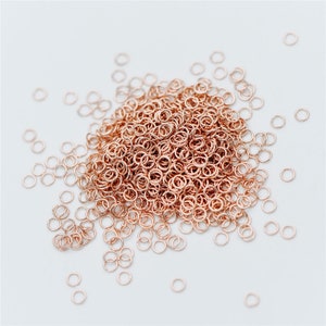 May include: A large pile of rose gold jump rings, scattered on a white surface. The rings are small and delicate, perfect for jewelry making.