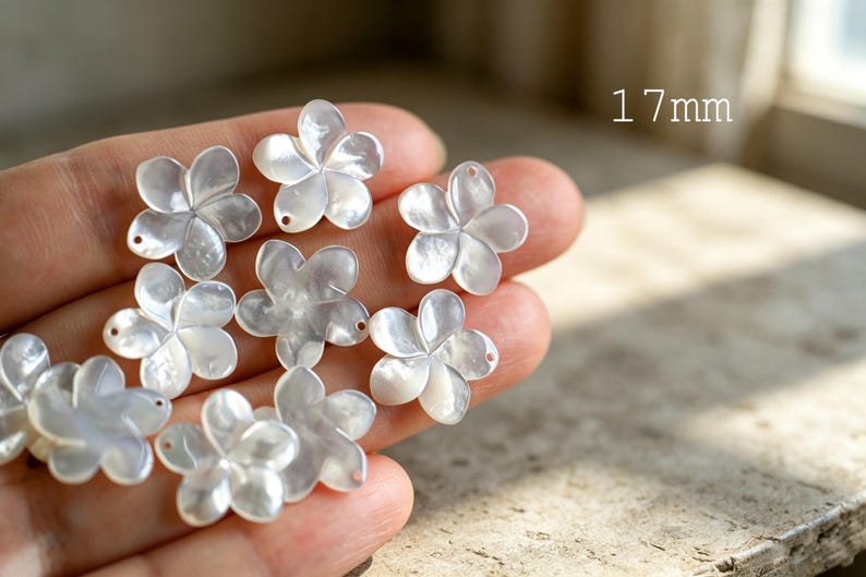 May include: A collection of white, iridescent, five-petaled flower-shaped charms made of mother of pearl. Each individual floral piece measures 17mm in diameter and features a small, circular hole near the edge for attachment purposes.