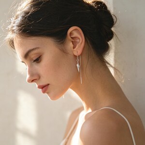 May include: A silver threader earring featuring a small, detailed seashell charm. The earring consists of a thin, flexible silver chain that passes through the earlobe, with the shell pendant hanging from one end of the delicate metal wire.