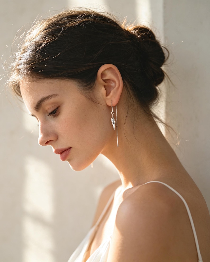 May include: A silver threader earring featuring a small, detailed seashell charm. The earring consists of a thin, flexible silver chain that passes through the earlobe, with the shell pendant hanging from one end of the delicate metal wire.