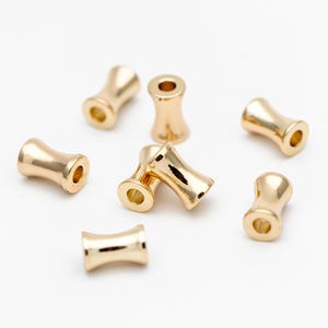 May include: Eight gold-colored metal beads with a cylindrical shape and a slightly wider middle section. The beads are scattered on a white surface.