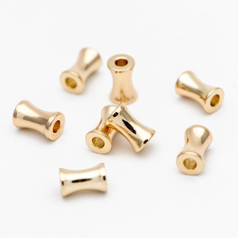 May include: Eight gold-colored metal beads with a cylindrical shape and a slightly wider middle section. The beads are scattered on a white surface.