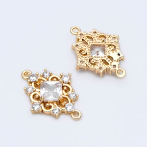 May include: Two gold-tone metal connector charms with clear crystal accents. Each charm features a square-shaped centre stone and intricate filigree details.