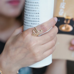 May include: A gold-coloured snake ring with a coiled design, worn on a finger. The ring features a detailed snake head. The person is holding a white cylindrical object and a book with Chinese text.