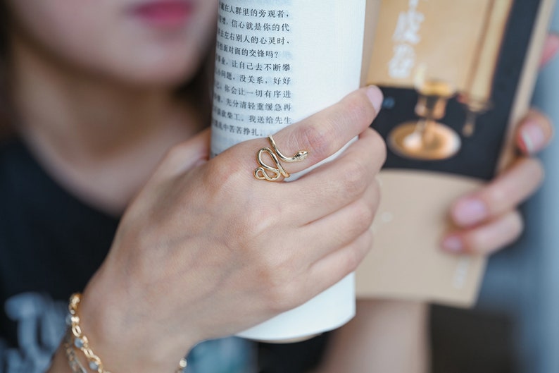 May include: A gold-coloured snake ring with a coiled design, worn on a finger. The ring features a detailed snake head. The person is holding a white cylindrical object and a book with Chinese text.