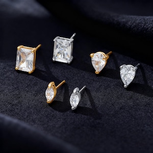 Peut inclure: Plusieurs boucles d'oreilles clous avec des pierres de zircone cubique transparentes. Les boucles d'oreilles présentent différentes formes, notamment rectangulaires, en poire et en marquise, serties dans des montures en or et en argent. Présentées sur un fond bleu foncé.