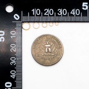 May include: A set of six gold jump rings of varying sizes, measured against a ruler and a US quarter. The jump rings are all round and have a smooth finish.