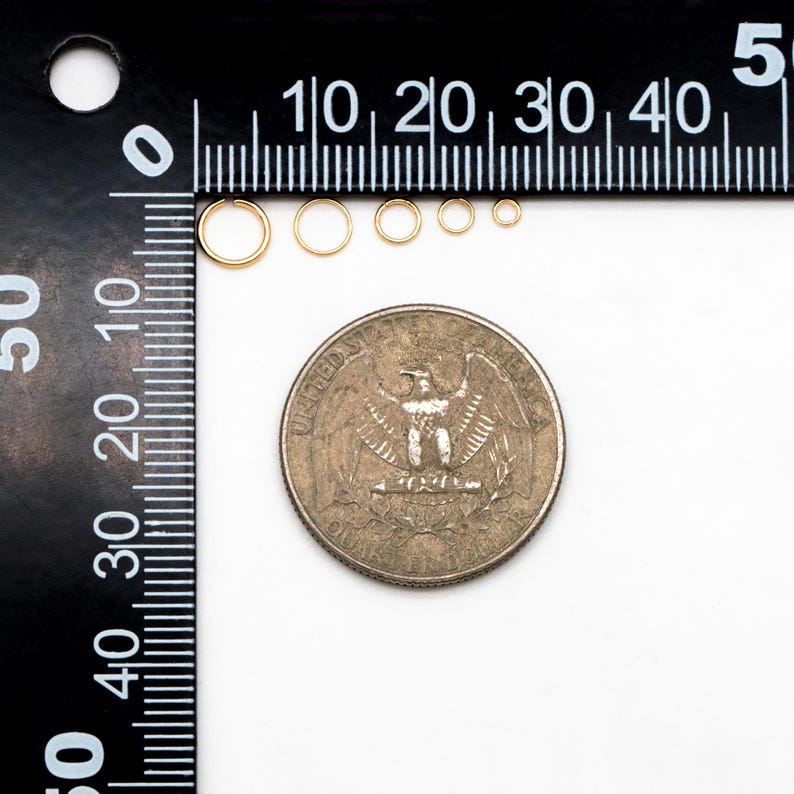May include: A set of six gold jump rings of varying sizes, measured against a ruler and a US quarter. The jump rings are all round and have a smooth finish.