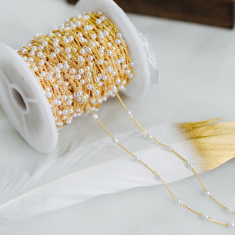 May include: A spool of gold chain with white pearl beads. The chain is unraveled and laid out on a white surface.