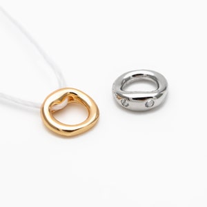 Könnte beinhalten: Zwei Ringe, einer goldfarben und einer silberfarben, vor weißem Hintergrund. Der goldfarbene Ring ist an einer weißen Schnur befestigt. Der silberfarbene Ring hat zwei kleine, klare Steine. Die Ringe haben ein glattes, abgerundetes Design.