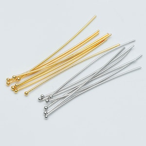 May include: A collection of gold and silver head pins with round ball ends. The pins are arranged in a fan shape on a white background.