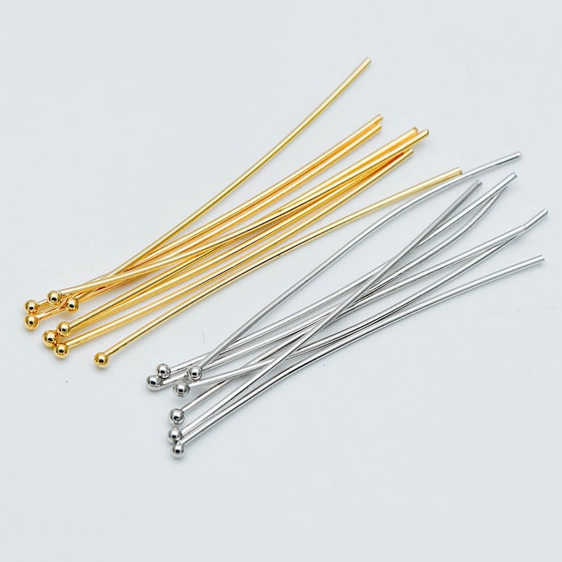 May include: A collection of gold and silver head pins with round ball ends. The pins are arranged in a fan shape on a white background.