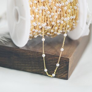 May include: A spool of gold chain with white pearl beads. The chain is on a wooden surface and a section of the chain is hanging down.