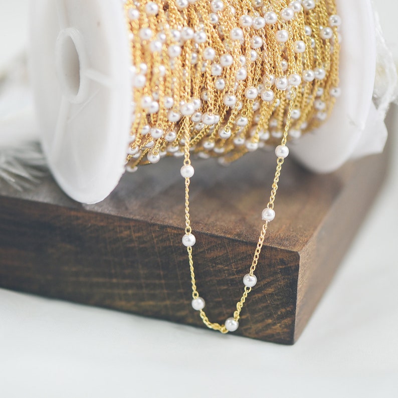 May include: A spool of gold chain with white pearl beads. The chain is on a wooden surface and a section of the chain is hanging down.