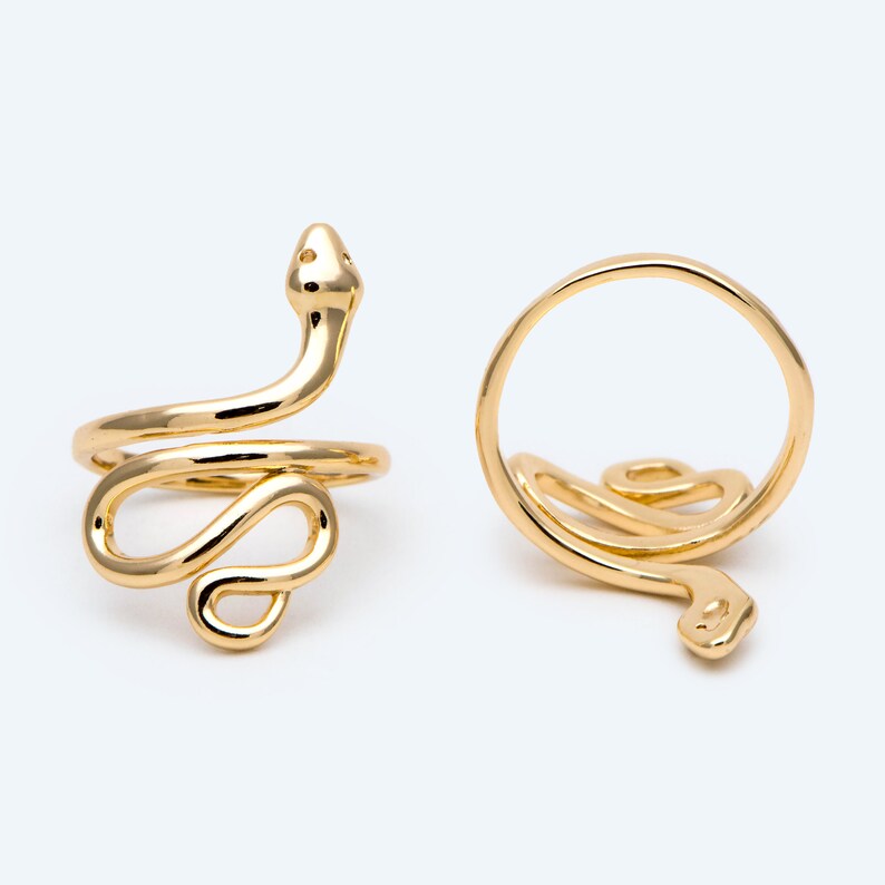 May include: Two gold-coloured snake rings. One ring is shown from the side, with the snake's head and body wrapping around the finger. The other ring is shown from above, with the snake's head and body forming a loop. The rings are on a white background.