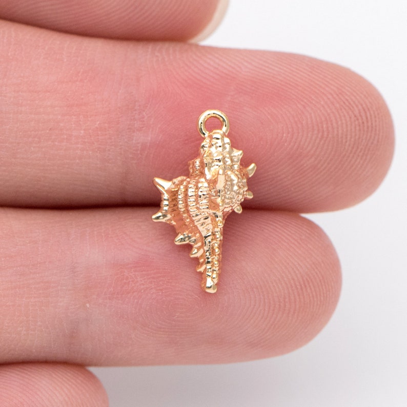 May include: Gold-toned seashell charm with a detailed, intricate design. The charm has a loop at the top for attaching to jewelry or other crafts.