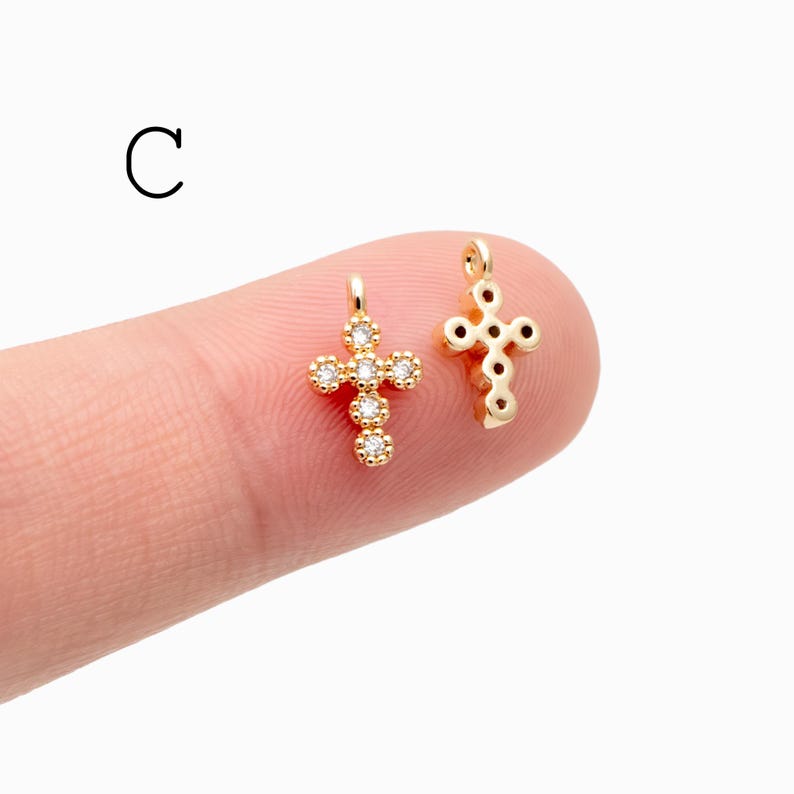 Pu&ograve; includere: Due piccoli ciondoli a forma di croce color oro. Una croce &egrave; adornata con pietre preziose rotonde e trasparenti. L'altra croce ha un design semplice e aperto. La lettera "C" si trova nell'angolo in alto a sinistra.