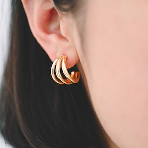 May include: Gold hoop earrings with three stacked rings. The earrings are worn in a person's ear.