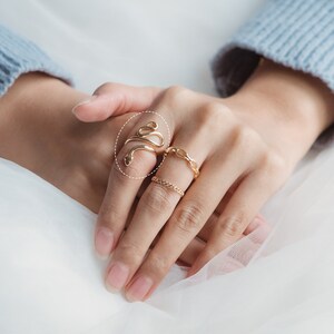 May include: A hand adorned with three gold rings. The standout ring features a snake design, spiraling around the finger. The other rings display a chain link and a small chain pattern. The background shows a light blue sweater.