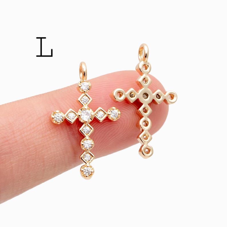 Pu&ograve; includere: Due ciondoli a forma di croce color oro con gemme chiare e scintillanti. Una croce &egrave; tenuta da un dito. Le croci hanno un anello nella parte superiore per essere appese. La lettera "L" &egrave; nell'angolo in alto a sinistra.