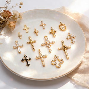 Peut inclure: Une collection de pendentifs croix dorés de différents modèles, dont certains ornés de pierres précieuses claires et noires, et de perles. Les croix sont présentées sur une assiette blanche légèrement texturée, avec des fleurs séchées en arrière-plan.