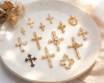 May include: A collection of gold-colored cross pendants with various designs, including those adorned with clear and black gemstones, and pearls. The crosses are displayed on a white, slightly textured plate, with dried flowers in the background.
