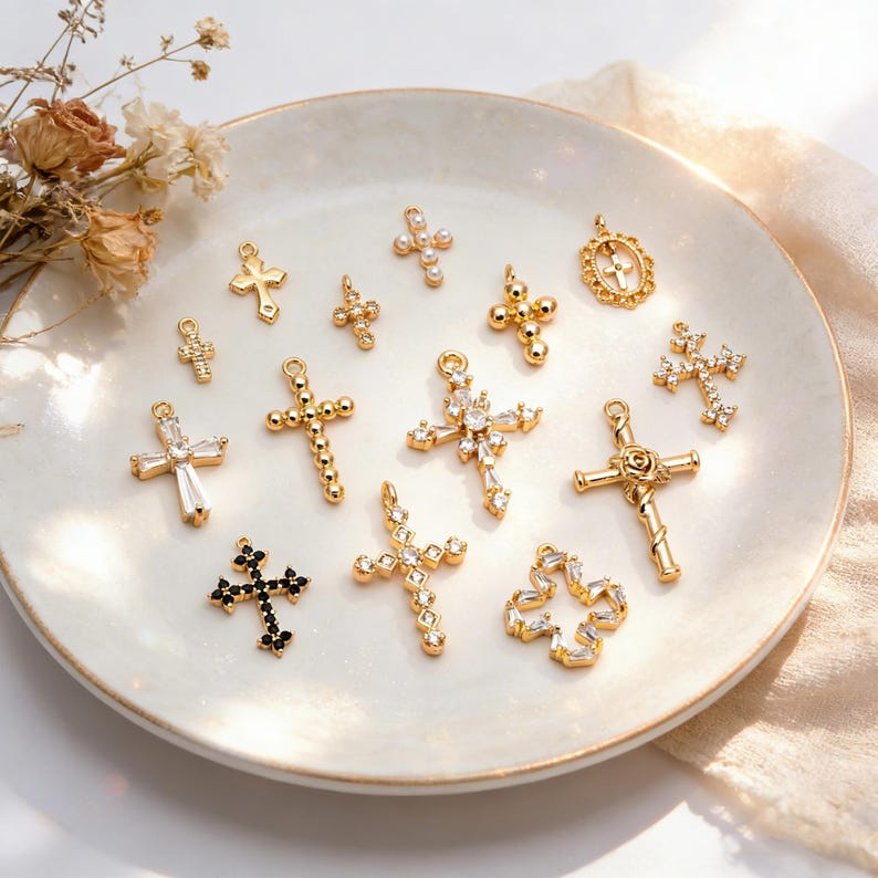 Pu&ograve; includere: Una collezione di ciondoli a forma di croce dorati con vari design, inclusi quelli adornati con pietre preziose chiare e nere e perle. Le croci sono esposte su un piatto bianco, leggermente strutturato, con fiori secchi sullo sfondo.
