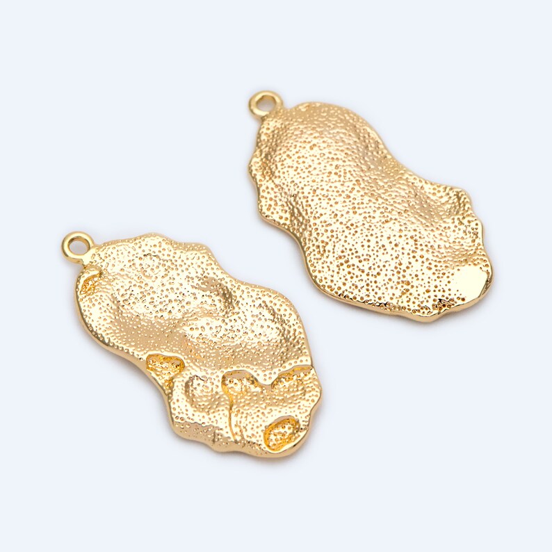 May include: Two gold-toned metal charms shaped like irregular ovals with a textured surface. Each charm has a small loop at the top for attaching to jewellery.