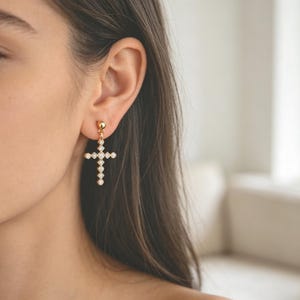 Pu&ograve; includere: Orecchini a forma di croce color oro con pietre trasparenti e scintillanti. Gli orecchini sono un simbolo religioso, con una piccola sfera d'oro in cima e una croce composta da pi&ugrave; pietre. Gli orecchini sono indossati sull'orecchio.