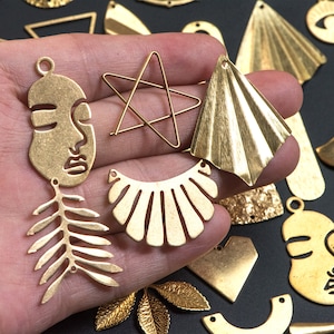 May include: Assortment of gold-toned metal charms in various shapes, including a face, a star, a leaf, a fan, and a scallop shell. The charms are displayed on a hand.