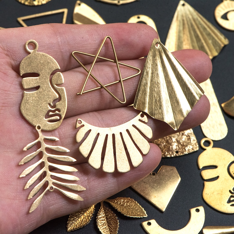 May include: Assortment of gold-toned metal charms in various shapes, including a face, a star, a leaf, a fan, and a scallop shell. The charms are displayed on a hand.
