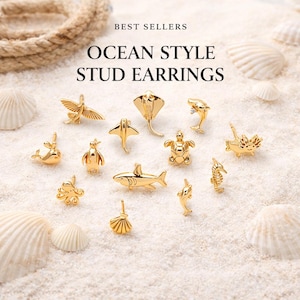 Può includere: Orecchini a lobo dorati a tema oceano, tra cui un cavalluccio marino, uno squalo e un delfino, esposti su una superficie sabbiosa. Il testo recita "BEST SELLERS" e "OCEAN STYLE STUD EARRINGS".