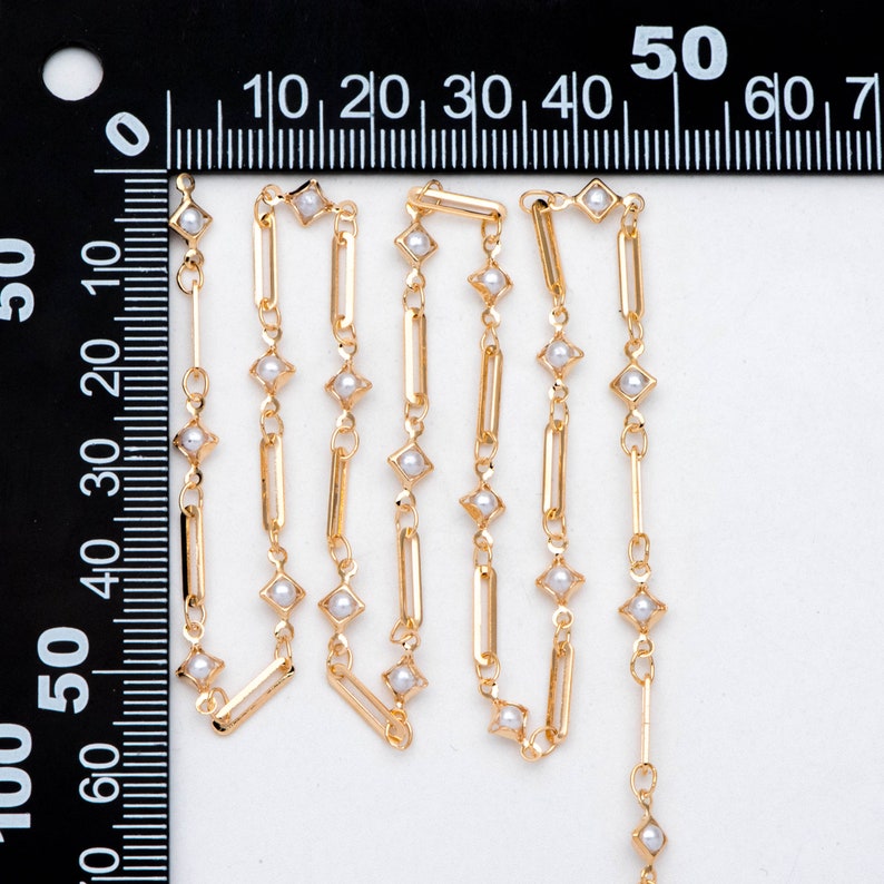 May include: Gold chain with small white pearl accents. The chain is made of rectangular and square links.