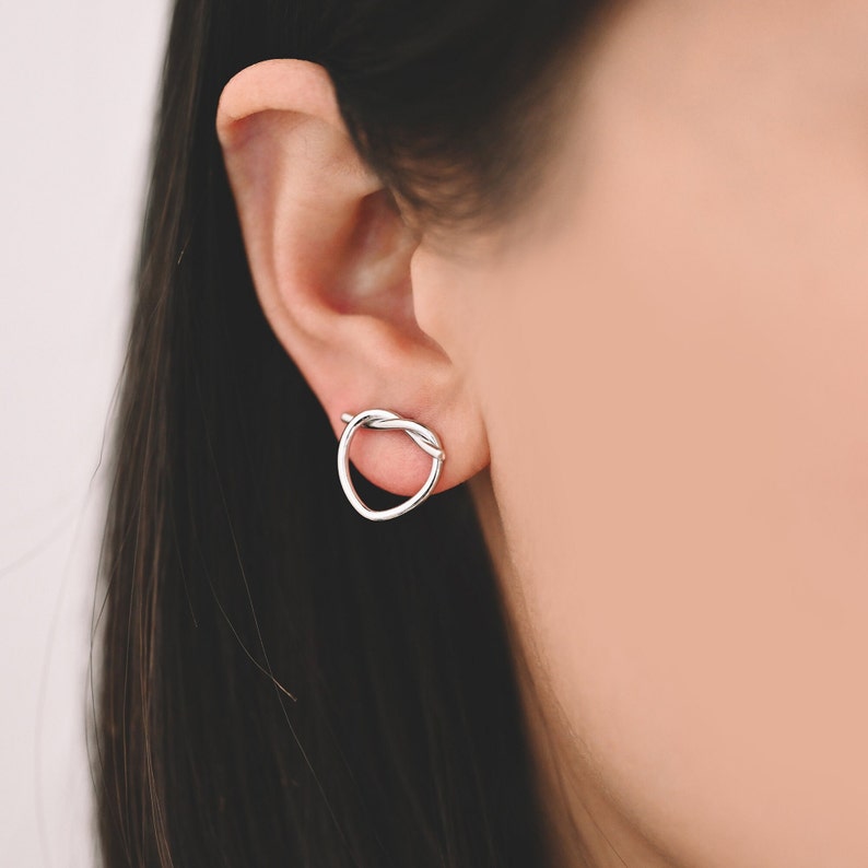 May include: Silver heart-shaped earrings with a twisted design. The earrings are shown on a model's ear.