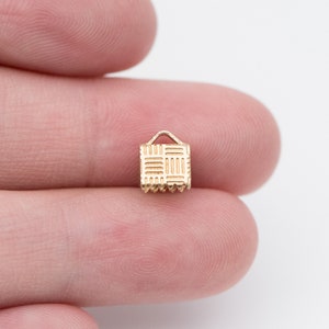 Puede incluir: Un peque&ntilde;o dije dorado con forma de cesta en miniatura. El dije tiene un patr&oacute;n tejido en la parte delantera y un peque&ntilde;o asa en la parte superior. El borde inferior tiene un dise&ntilde;o festoneado. El dije se sostiene entre dos dedos.