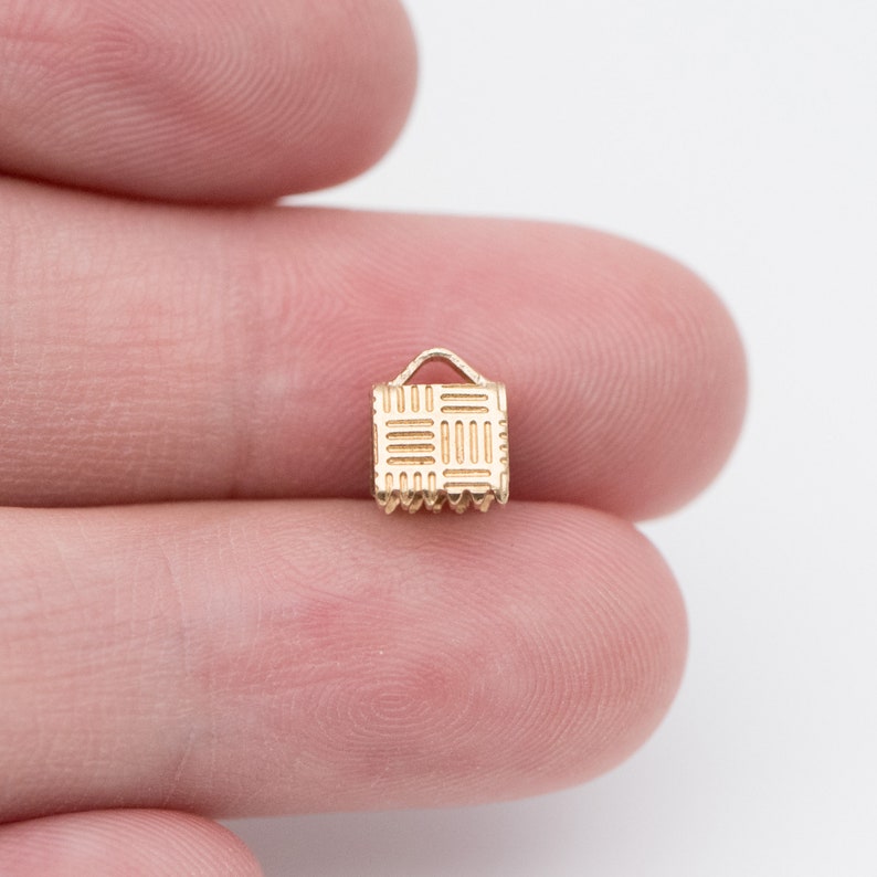 Puede incluir: Un peque&ntilde;o dije dorado con forma de cesta en miniatura. El dije tiene un patr&oacute;n tejido en la parte delantera y un peque&ntilde;o asa en la parte superior. El borde inferior tiene un dise&ntilde;o festoneado. El dije se sostiene entre dos dedos.