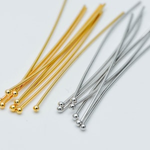 May include: A close-up of two bundles of head pins, one gold and one silver. The head pins are arranged in a fan shape, with the heads of the pins facing the viewer. The pins are about 2 inches long.