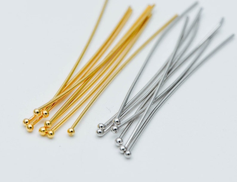May include: A close-up of two bundles of head pins, one gold and one silver. The head pins are arranged in a fan shape, with the heads of the pins facing the viewer. The pins are about 2 inches long.