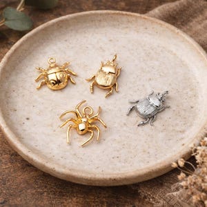 Pode incluir: Quatro pingentes com tema de insetos em tons de ouro e prata. Os pingentes incluem uma joaninha, um besouro, uma aranha e um besouro escaravelho. Os pingentes são exibidos em um prato de cerâmica bege. Ideal para fazer joias ou artesanato.